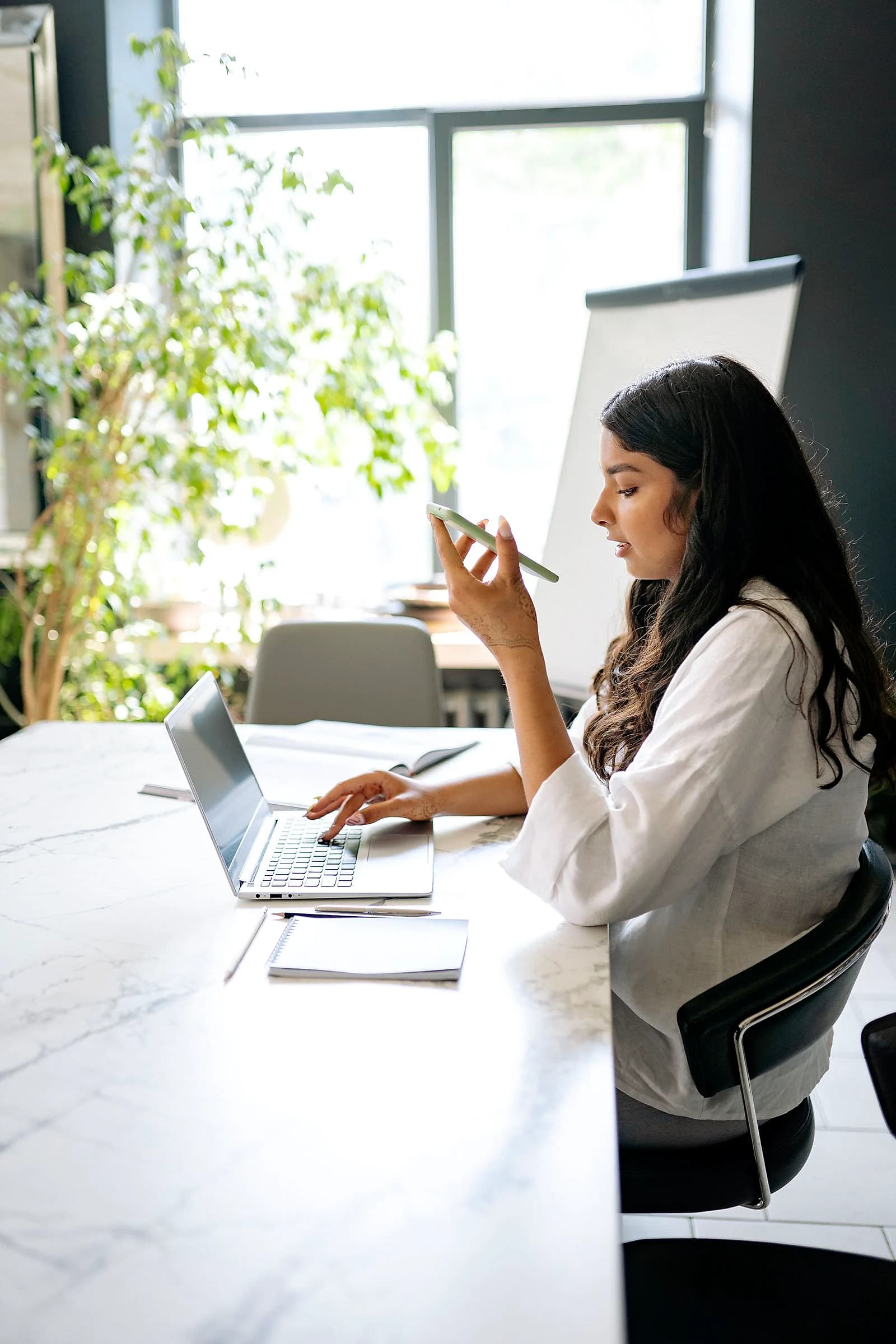 A woman at an office desk types on a laptop while taking a call